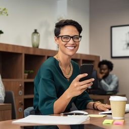 Happy fashionable girl using smartphone while working on desktop computer. Cheerful young business woman sitting in coworking space  and looking at camera. Portrait of  freelancer with eyeglasses.