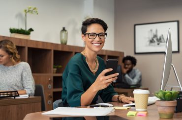 Happy fashionable girl using smartphone while working on desktop computer. Cheerful young business woman sitting in coworking space  and looking at camera. Portrait of  freelancer with eyeglasses.